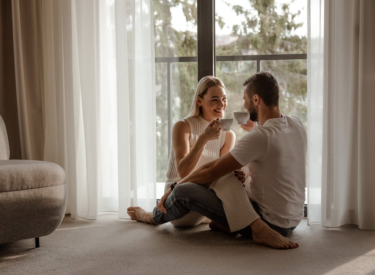 Couple enjoying coffee by a large window with balcony, sheer curtains, soft carpet and cozy seating.