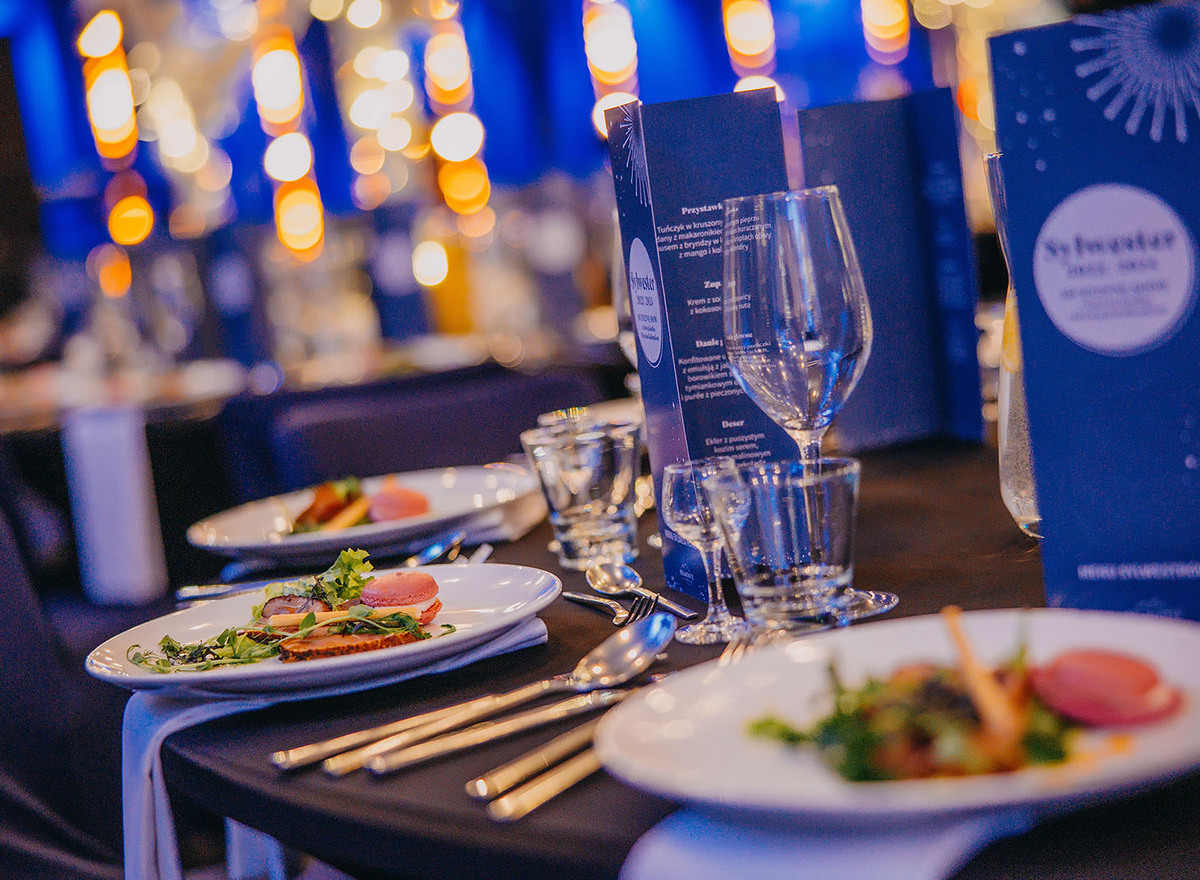 Elegant banquet table with plated appetizers, wine glasses, menus and warm blurred lights in a blue-lit dining room