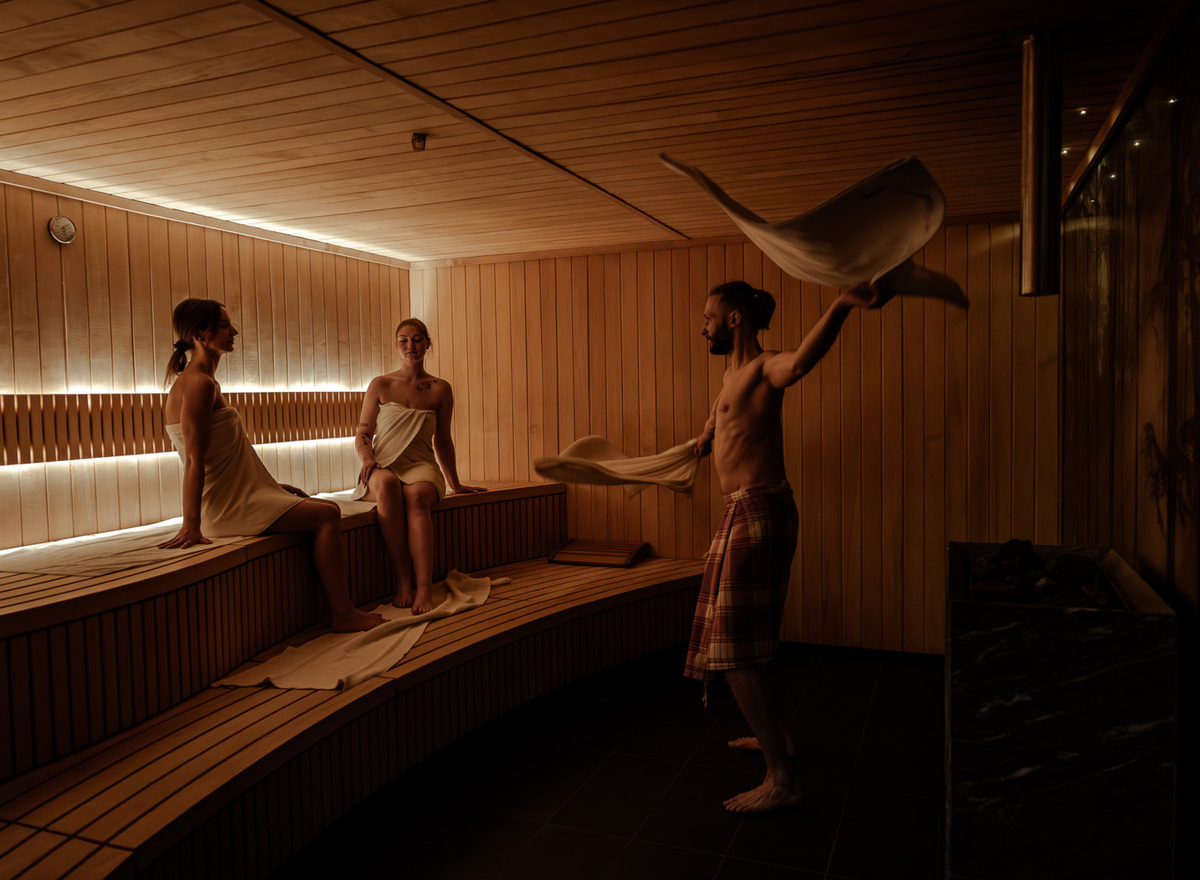 Drewniana sauna w hotelu, dwie kobiety siedzą na ławkach, mężczyzna wachluje ręcznikiem podczas relaksującego seansu
