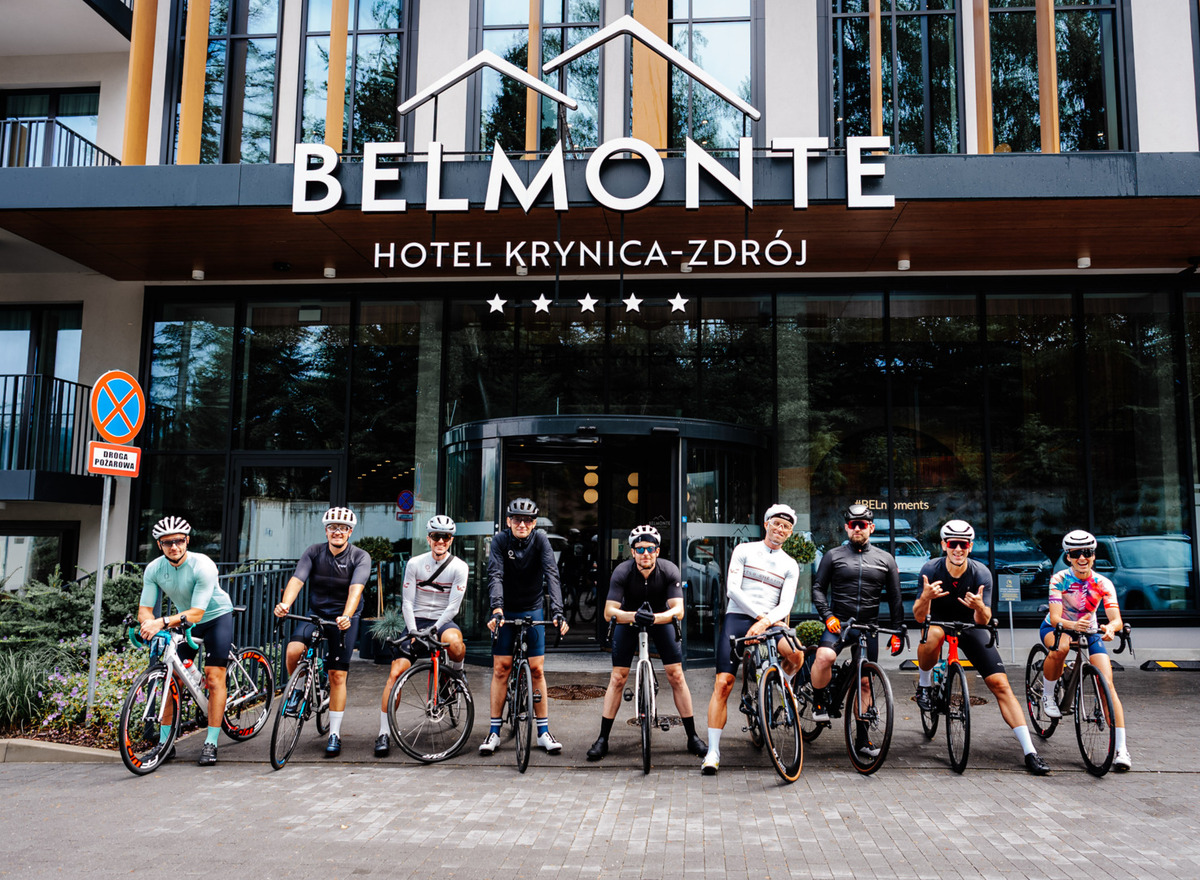 Deväť cyklistov na cestných bicykloch pózuje pred moderným hotelom Belmonte Krynica‑Zdrój pod päťhviezdičkovým nápisom.