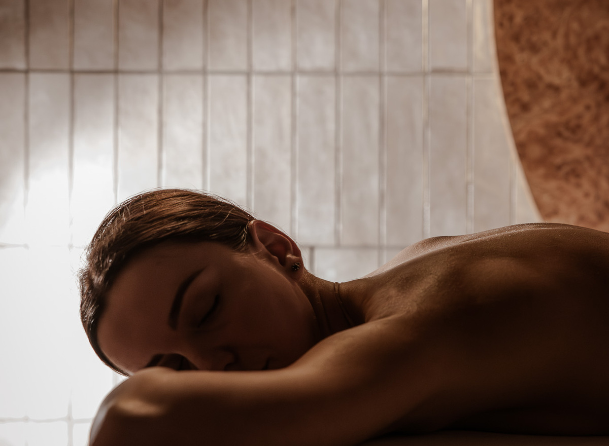 Guest lying face down on a massage table, skin glistening under warm spa light, tiled wall and curved wooden accent in the room