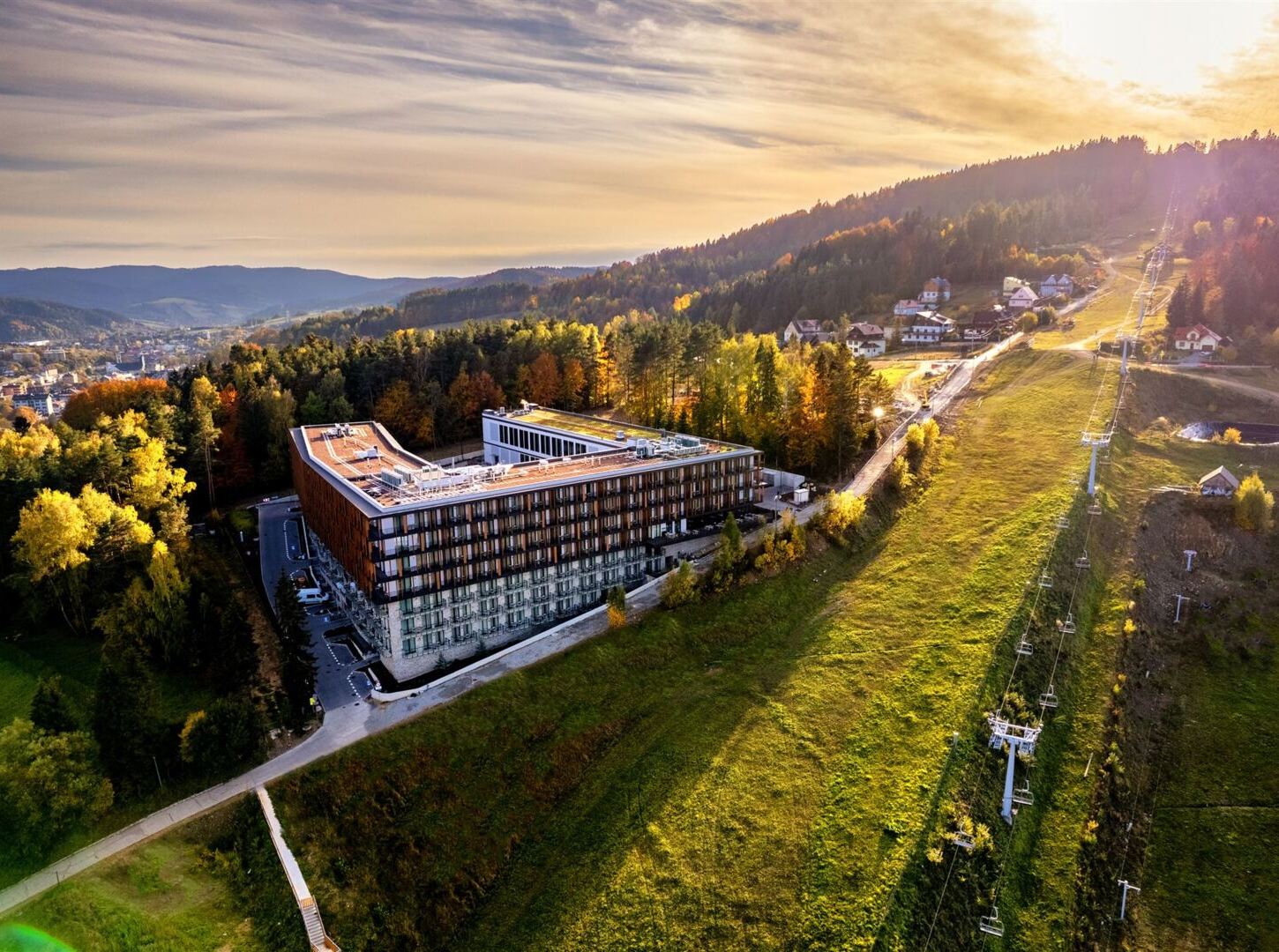 Nowoczesny górski hotel z balkonami przy stoku narciarskim i wyciągu, jesienny las i widok na dolinę o zachodzie słońca