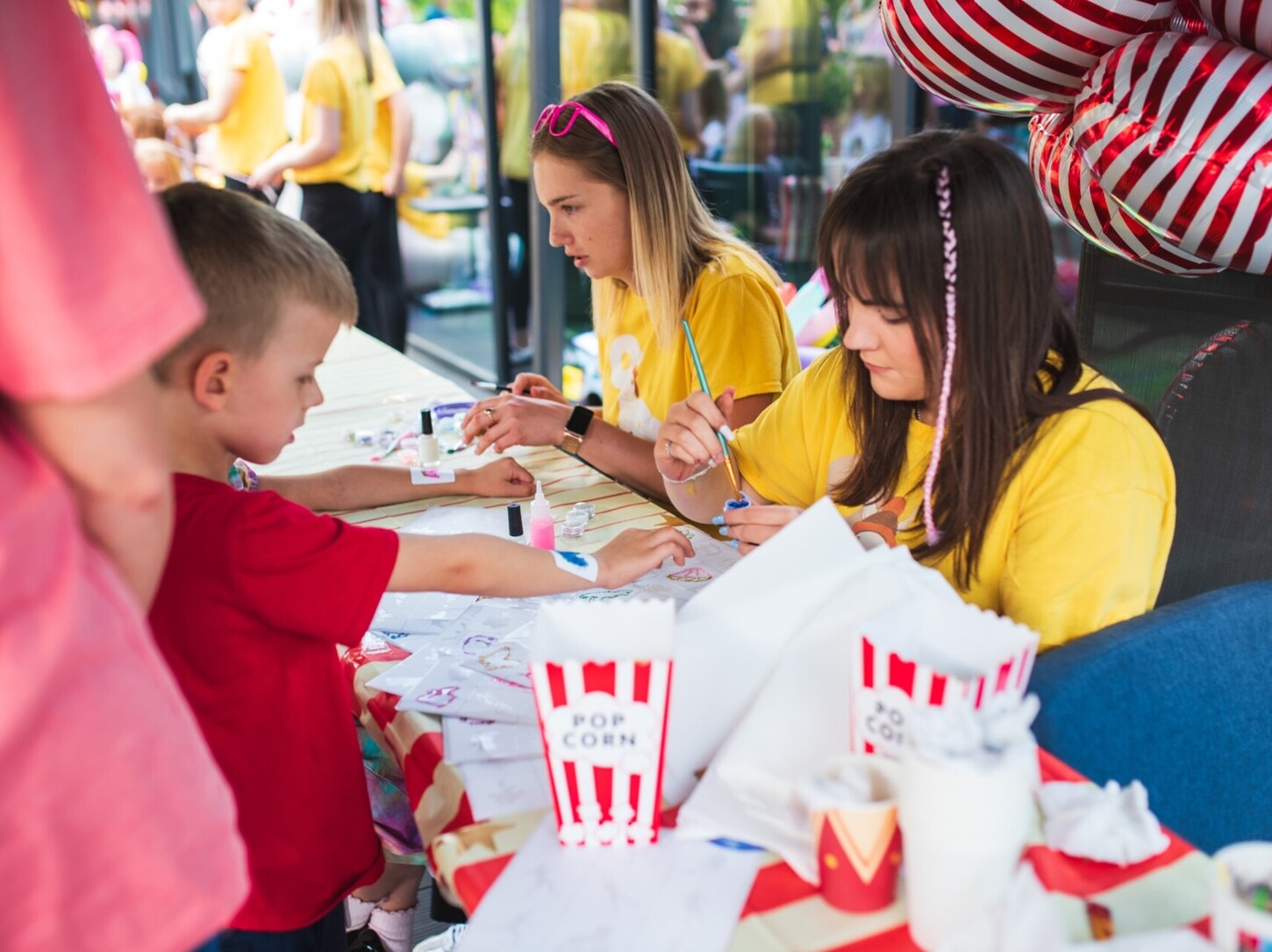Animatorki klubu dziecięcego malują na rękach przy stole plastycznym, popcorn i czerwono-białe balony, strefa przyjazna rodzinom.