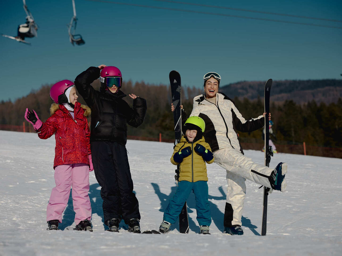 Family in colorful ski gear laughing on a sunny snowy slope, skis upright and chairlift passing overhead