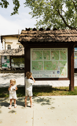 Drewniany, zadaszony punkt informacyjny z mapami tras MTB pod drzewami; dwoje dzieci ogląda mapy przy zabytkowym budynku uzdrowiska.