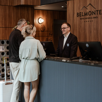 Pár sa registruje na recepcii Hotela Belmonte — usmievavý recepčný, batožina, drevené panely a teplé osvetlenie