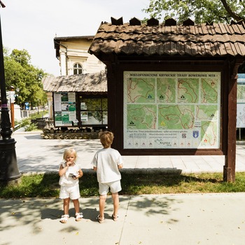 Drevený informačný stánok s mapami MTB trás v tienistom parku; dve deti, jedno líže zmrzlinu.