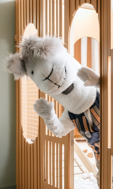 Fluffy white mascot peeks through warm wooden slats into a hotel's kids' playroom with slide and white ball pit