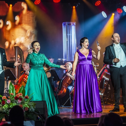 Four opera singers in formal eveningwear toast with champagne on stage during a gala concert, orchestra and floral arrangements.