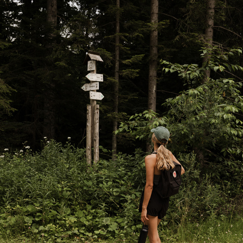 Woman with backpack and water bottle on a forest trail reading a wooden signpost beside tall pine trees