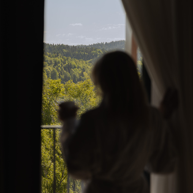 Hosť na hotelovom balkóne v župane drží hrnček, medzi závesmi je výhľad na slnečné zalesnené kopce.