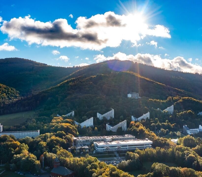 Complesso alberghiero moderno immerso in colline verdi con vista sulle montagne sotto un cielo azzurro luminoso.