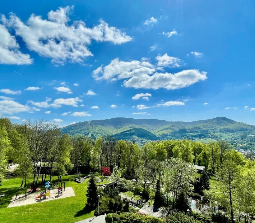 Grön park med lekplats omgiven av träd och kullar under blå himmel med moln.