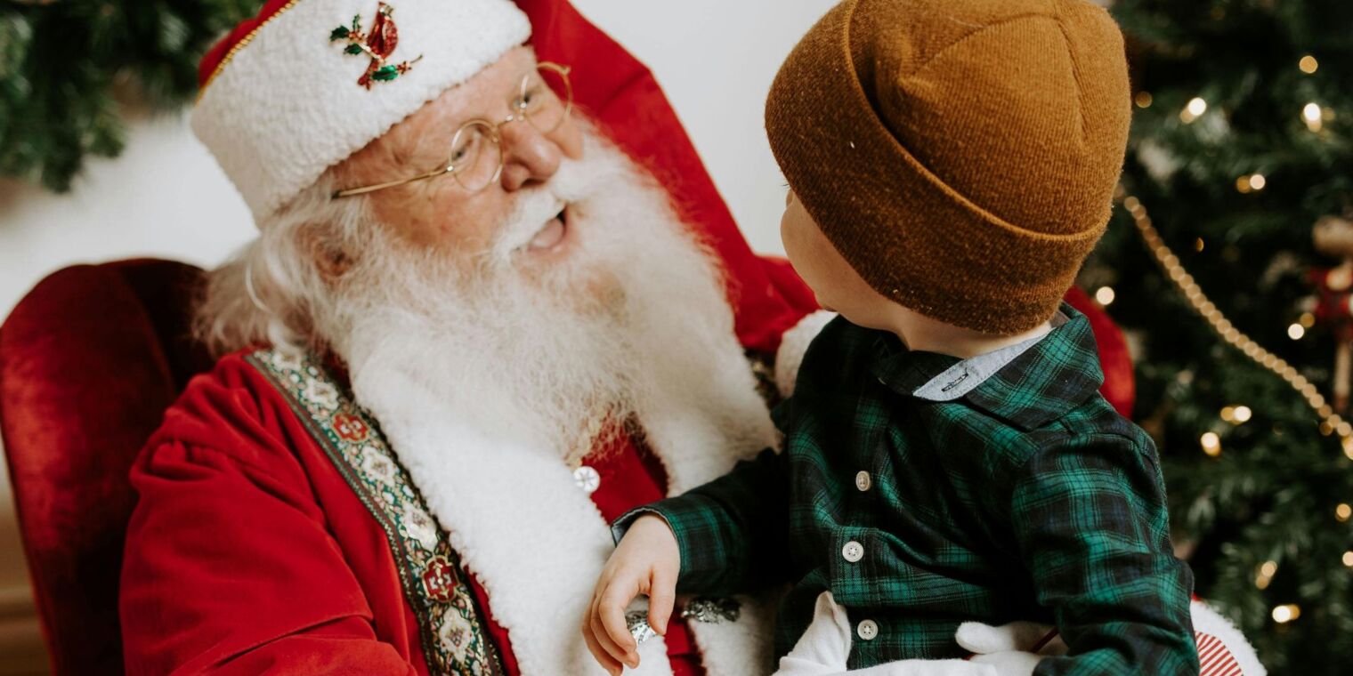Wesoły Święty Mikołaj rozmawia z małym chłopcem przy udekorowanej choince w świątecznym wnętrzu hotelu.
