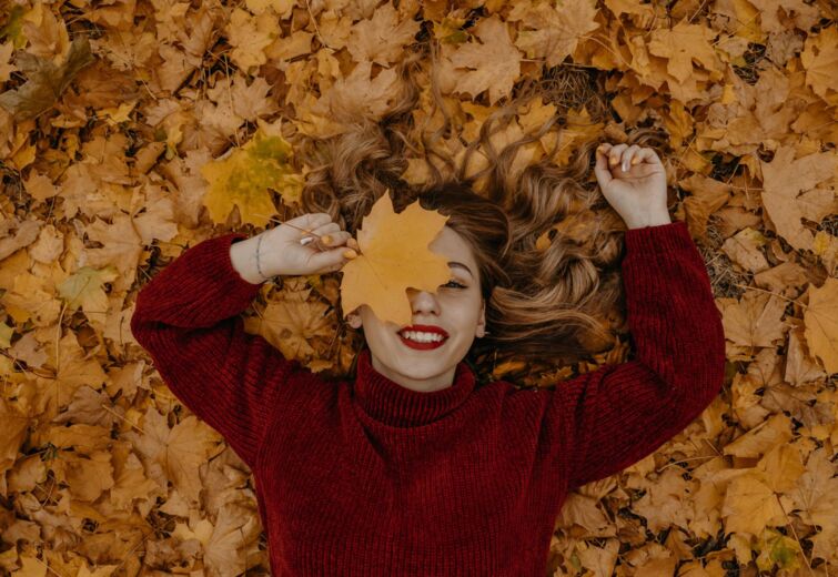 Young woman in a burgundy sweater lying on a bed of autumn leaves, covering one eye with a maple leaf.