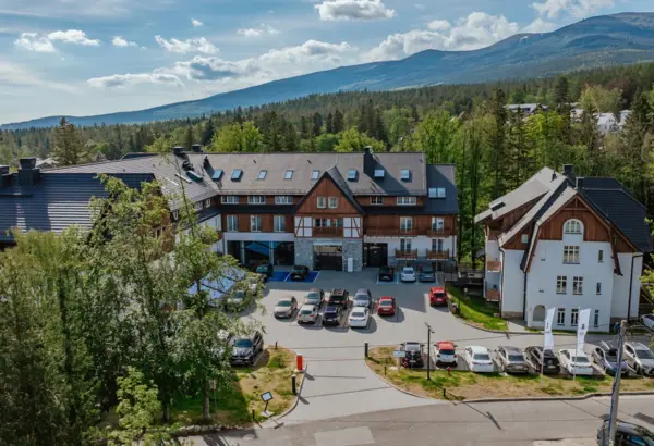 Hotel górski z tradycyjną drewnianą architekturą, parkingiem i widokiem na zalesione wzgórza i góry w tle.