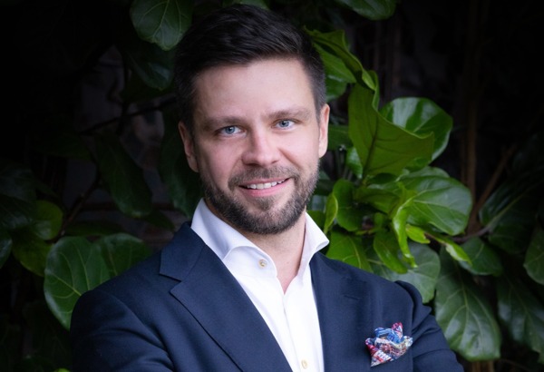 Muž v tmavě modrém obleku a bílé košili se usmívá, má založené ruce, za ním jsou zelené listy