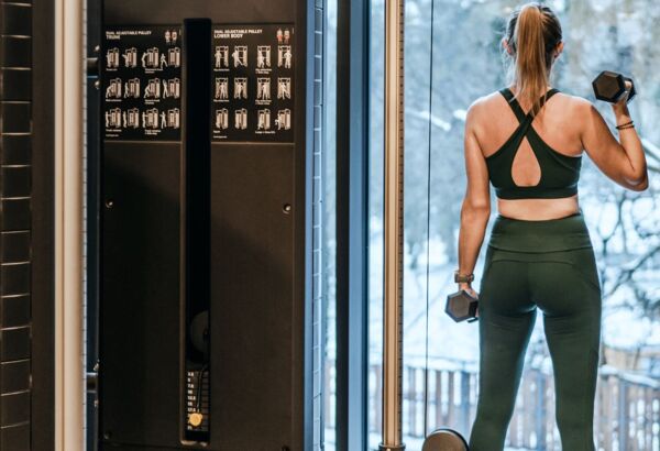 Gość w siłowni hotelowej ćwiczy z hantlami przy dużym oknie z widokiem na zimowy ogród na zewnątrz.