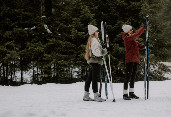 Dwie kobiety w zimowych ubraniach trzymają narty stojąc na zaśnieżonym terenie przy lesie iglastym.