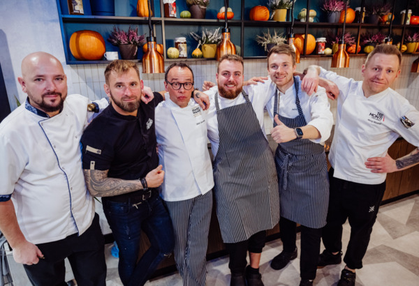 Team of six chefs in a restaurant kitchen with pumpkin and plant decorations, wearing aprons and white chef jackets.