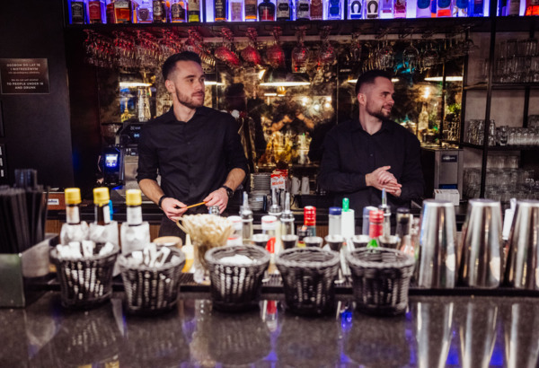 Two bartenders in black shirts stand behind a well-stocked bar with hanging glasses and bottles of alcohol