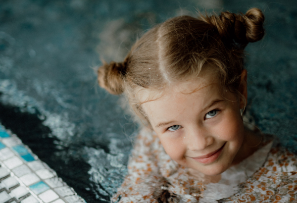 Smiling girl with two buns swimming and holding the edge of an indoor pool with mosaic tile flooring