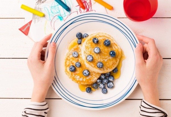 Two fluffy pancakes topped with syrup and fresh blueberries on a plate, with colorful crayons and a red cup nearby