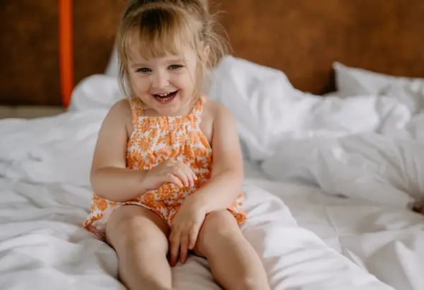Small child in summer dress sitting and smiling on a large bed in a bright hotel room