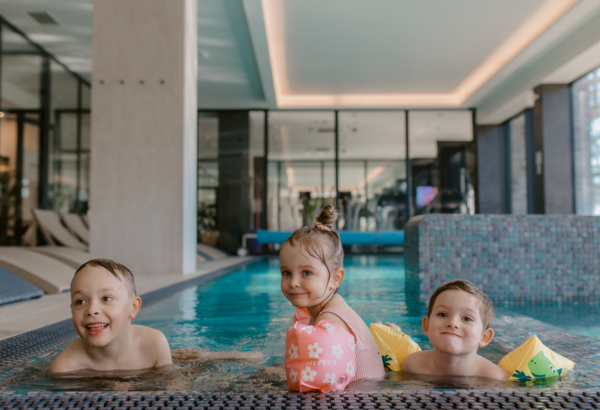 Drei Kinder spielen im modernen Innenpool eines Hotels mit Mosaikwänden und Liegestühlen im Hintergrund.