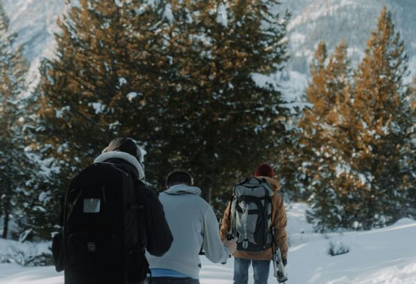 Trzech turystów z plecakami idzie zimową trasą przez zaśnieżony las iglasty w górskiej scenerii.