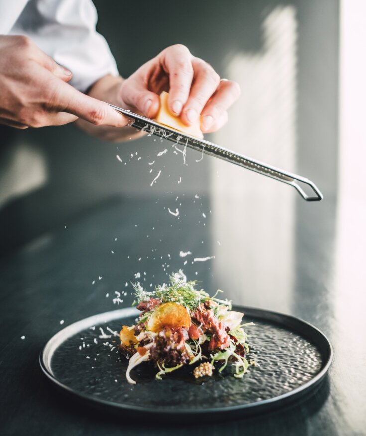 Šéfkuchař strouhá sýr na čerstvý bylinkový salát umělecky naaranžovaný na černém talíři v moderní restauraci.