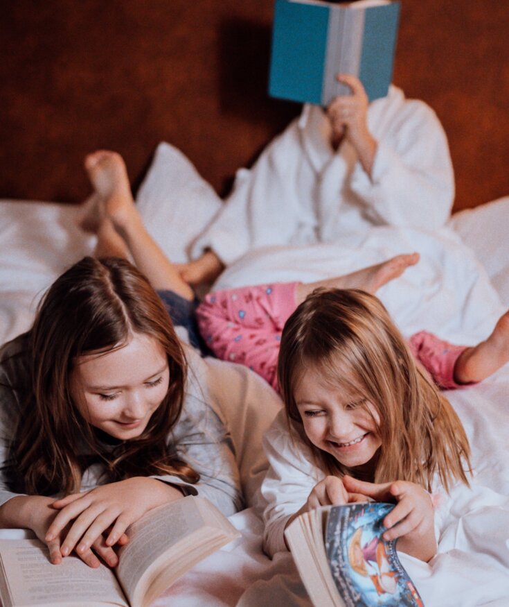 Děti čtou knihy na pohodlné hotelové posteli, v pozadí relaxuje osoba v bílém županu