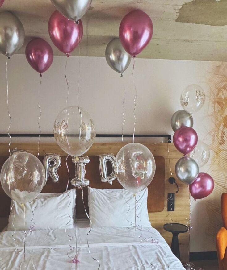 Przytulny pokój hotelowy udekorowany balonami i kwiatami na przyjęcie panieńskie z wygodnym łóżkiem i pomarańczowym fotelem.