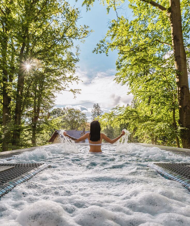 Gość relaksuje się w jacuzzi na świeżym powietrzu otoczonym bujną zielenią i promieniami słońca w rustykalnym otoczeniu.