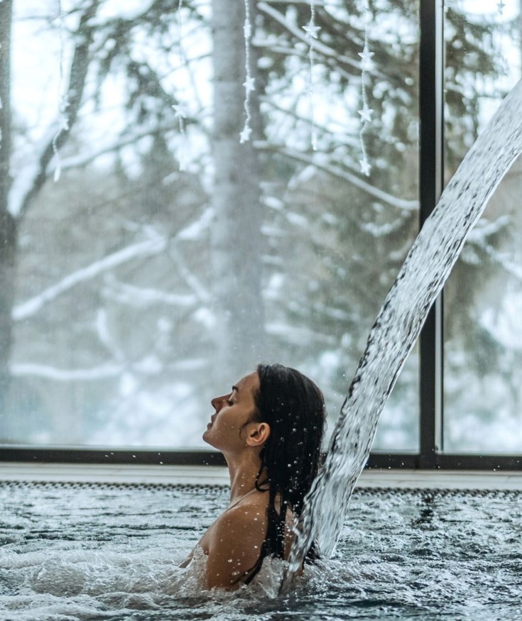 Gość relaksuje się pod strumieniem wody w krytym basenie z widokiem na ośnieżoną zimową scenerię za dużymi oknami