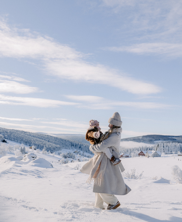 Woman in warm coat and hat holding child dressed for winter in snowy landscape with forested hills