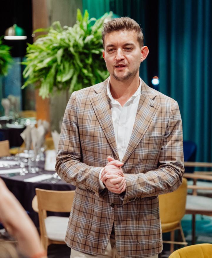 Man in beige plaid blazer talking in a restaurant with lush green plants and elegantly set tables