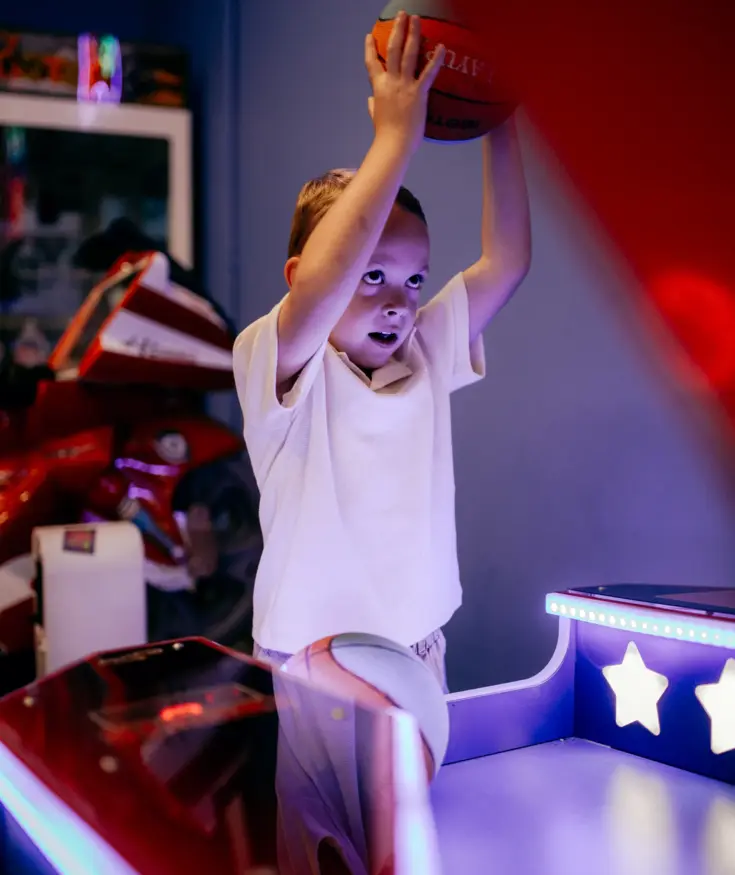 Child playing basketball in a brightly lit arcade room with neon stars and colorful gaming machines