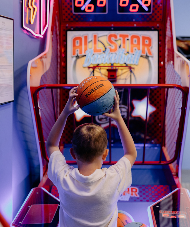 Child playing basketball arcade game in hotel game room with colorful neon lights and multiple basketballs ready to shoot.