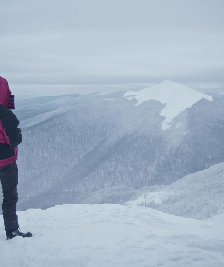Osoba w czerwono-czarnej kurtce podziwiająca zimowy górski krajobraz z białym szczytem i śniegiem wokół