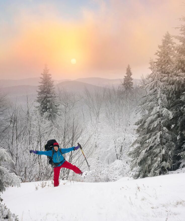 Osoba w czerwonym i niebieskim ubraniu cieszy się zimową wędrówką w śnieżnym lesie z ośnieżonymi drzewami i widokiem gór w tle