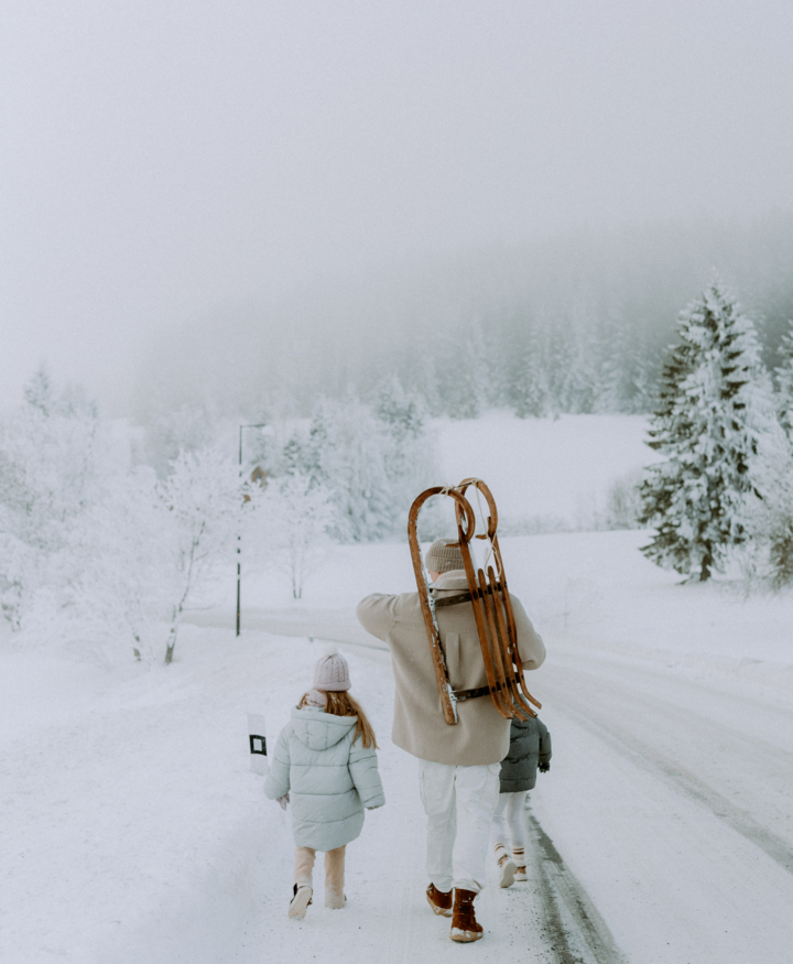 Family walking along snowy mountain road carrying wooden sleds, ready for winter fun and outdoor adventure.