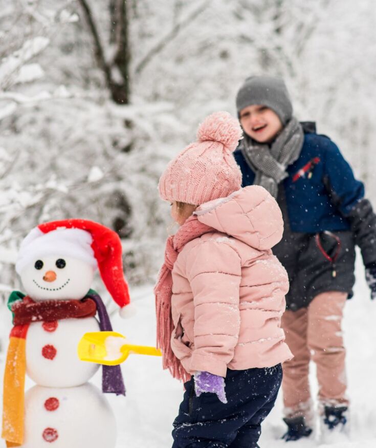 Zwei Kinder spielen mit einem Schneemann im verschneiten Wald, eines hält eine gelbe Schaufel