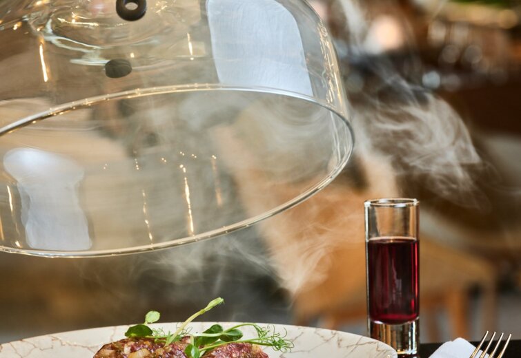 Gurmánské masové jídlo se zelenými bylinkami na elegantním mramorovém talíři pod skleněnou pokličkou, vedle sklenice červeného vína