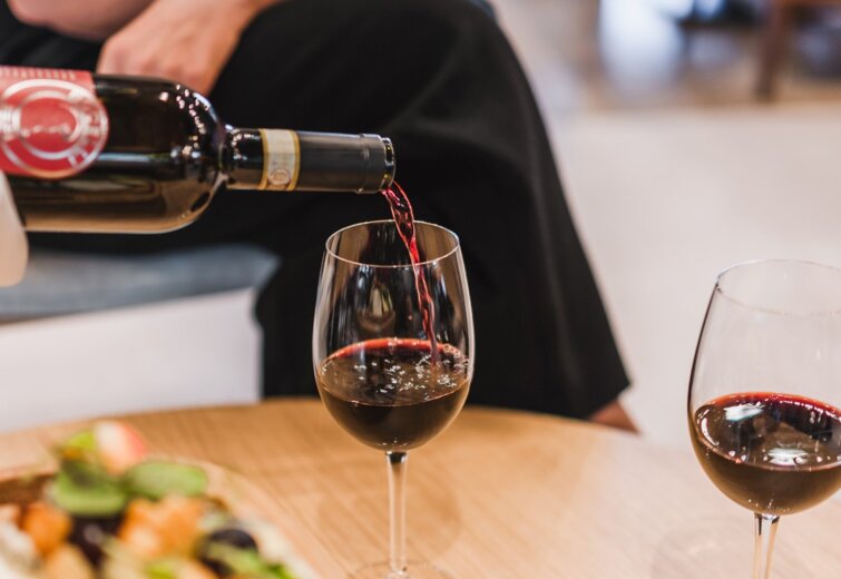 Osoba nalévá červené víno do sklenice v útulném salonku s občerstvením na dřevěném stole.