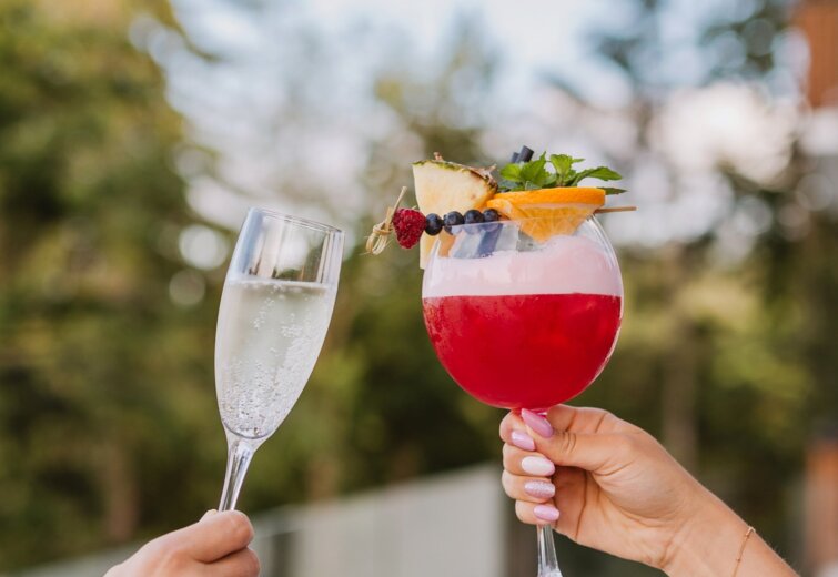 Dva lidé připíjejí sklenicemi s perlivým vínem a ovocným koktejlem venku s výhledem na zahradu.