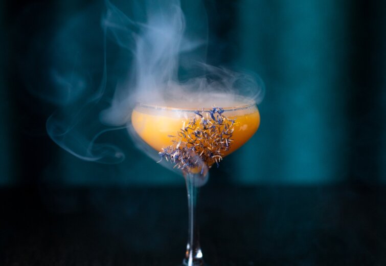 Elegantní oranžový koktejl ve sklenici s jemným kouřem a sušenými květy jako ozdobou
