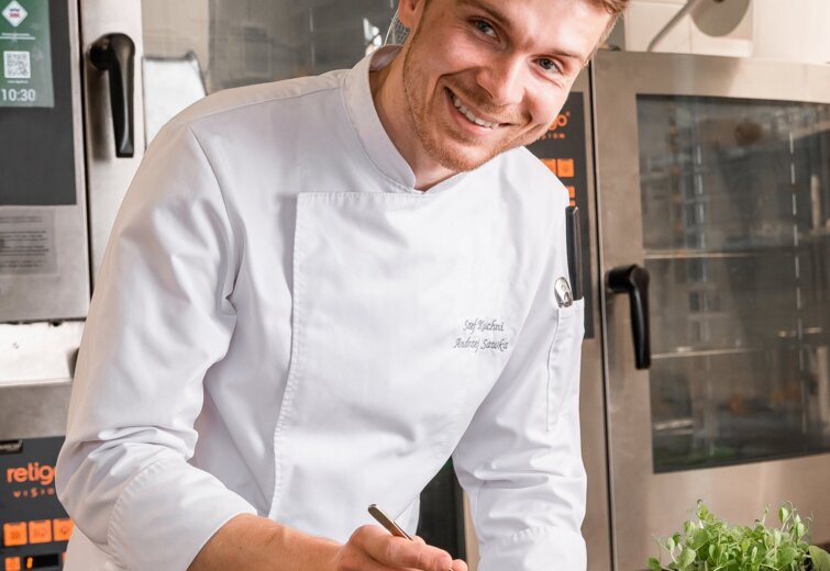 Usměvavý kuchař v bílém rouchu zdobí těstovinové jídlo čerstvými bylinkami v moderní hotelové kuchyni.