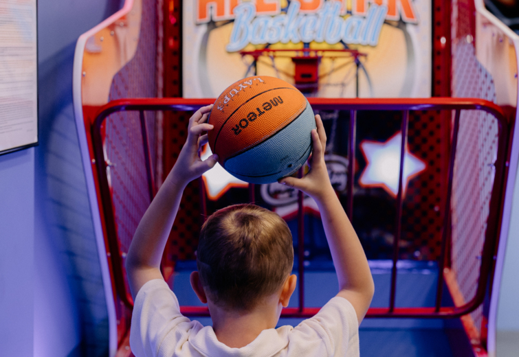 Kind im weißen Hemd spielt Basketball-Automat im Hotelspielzimmer mit bunten Lichtern.