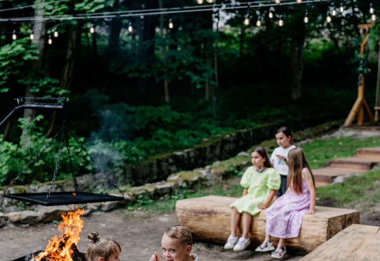 Children sitting on wooden benches around a campfire in a forest setting with string lights overhead.